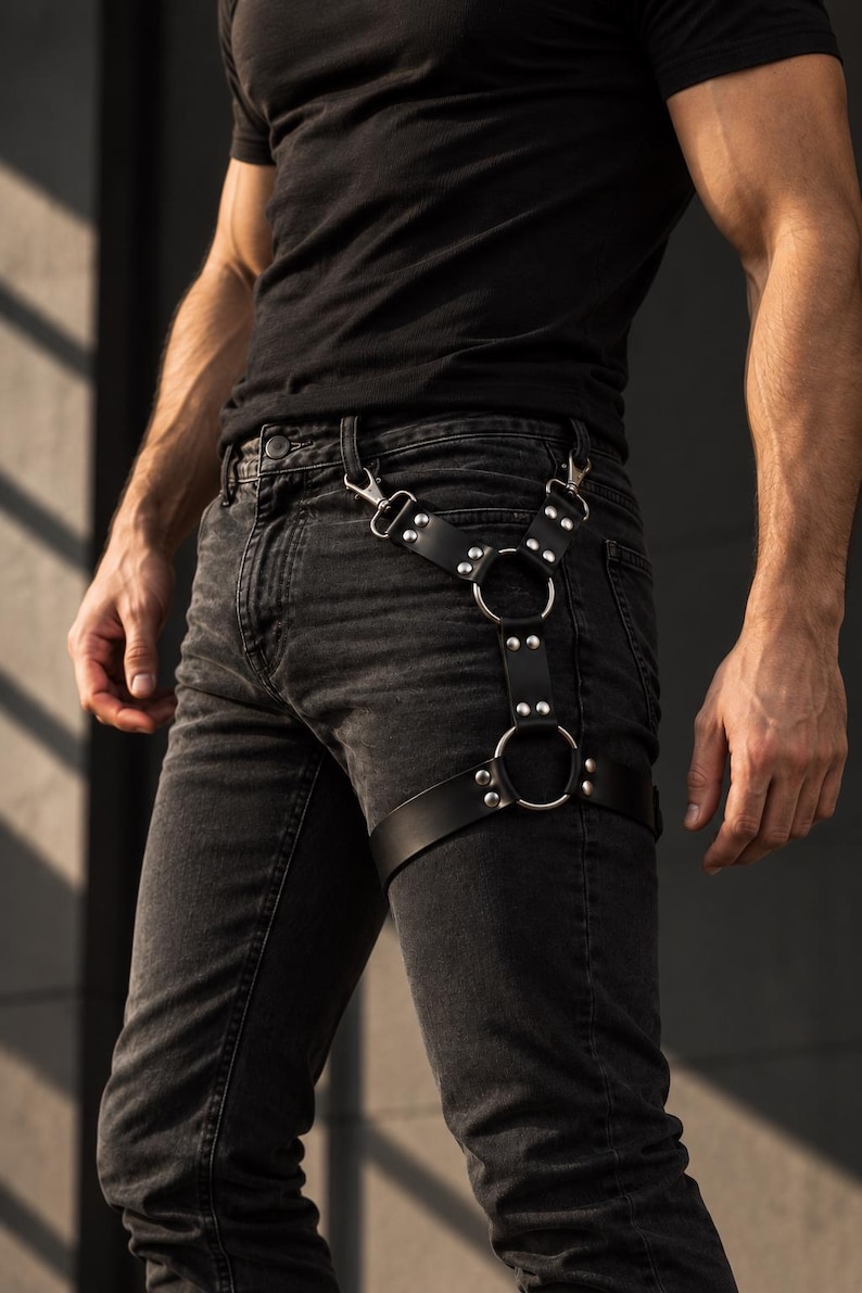 May include: A black leather leg harness featuring two silver-toned metal rings connected by adjustable straps with silver rivets. The harness attaches to belt loops with two metal clips and secures around the thigh with a wide leather strap.