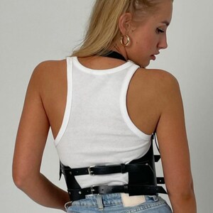 May include: A black leather corset with multiple straps and buckles. The corset is worn over a white tank top and blue jeans.