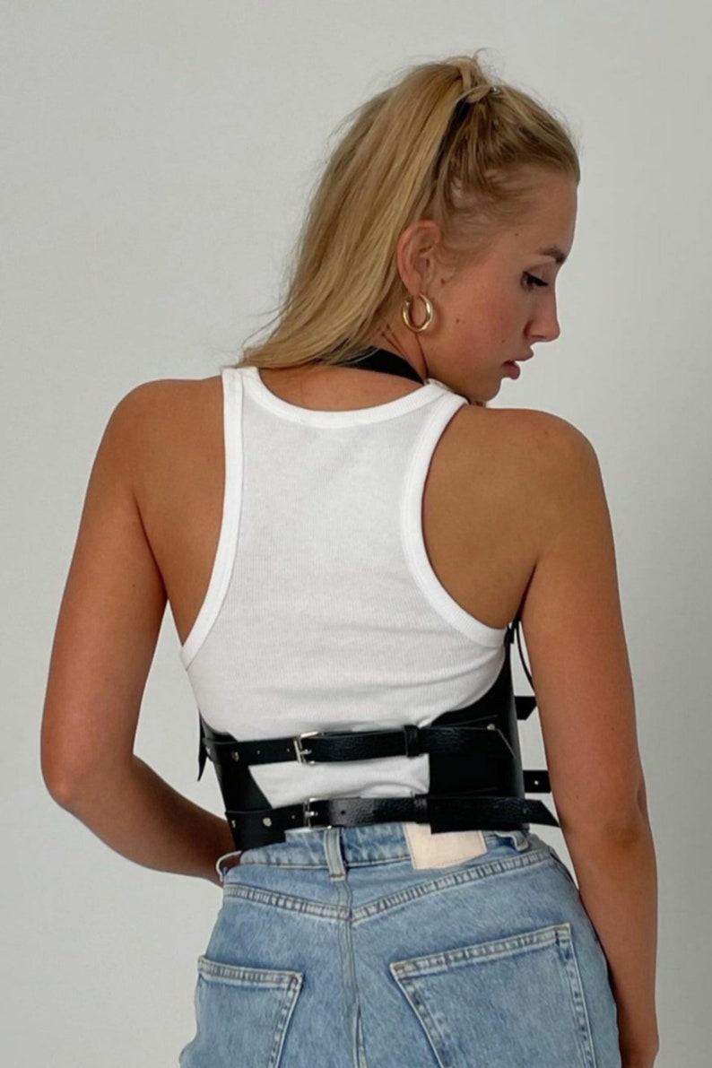 May include: A black leather corset with multiple straps and buckles. The corset is worn over a white tank top and blue jeans.