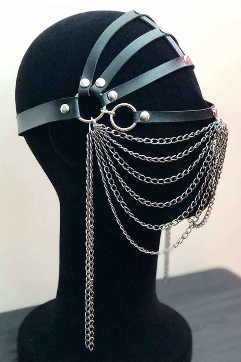 May include: Black leather face mask with a silver chain detail. The mask has a geometric design with multiple straps and studs.