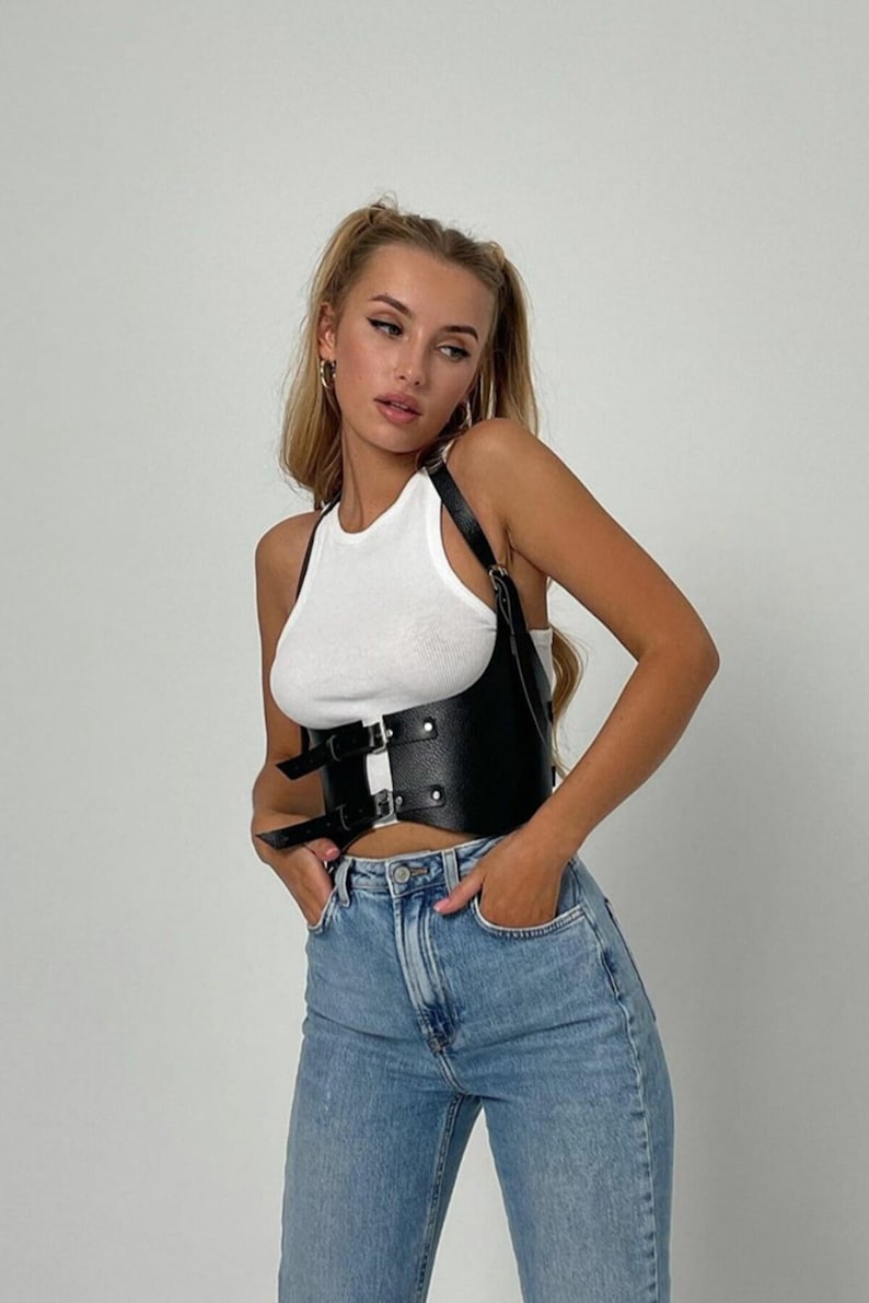 May include: A black leather corset with two buckles and adjustable straps. The corset is worn over a white tank top and blue jeans.