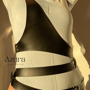 Puede incluir: Arn&eacute;s de cuero negro con una correa diagonal en el pecho y m&uacute;ltiples correas en la cintura. Se lleva sobre un body azul claro. El logotipo de Azura Exclusive es visible en la esquina inferior izquierda.