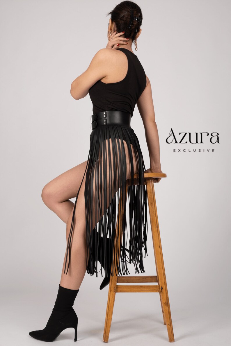 May include: Black leather fringe skirt with a wide belt and double buckle closure. The skirt is paired with a black top and black heeled boots. The Azura Exclusive logo is visible on the right side.