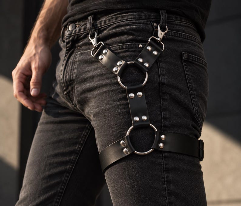 May include: A black leather leg harness featuring silver-toned metal rings and rivets. The accessory attaches to belt loops with two swivel clips and includes a horizontal strap that encircles the thigh, secured with additional metal hardware.