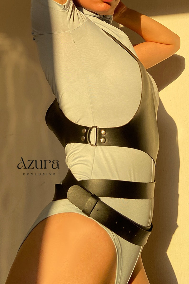 Puede incluir: Arn&eacute;s de cuerpo de cuero negro con hebilla y detalle de cintur&oacute;n, usado sobre un body gris claro. La imagen incluye el texto "AZURA EXCLUSIVE".