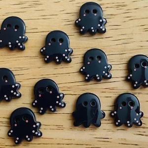 Puede incluir: Una colección de diez botones azules oscuros con forma de pulpo. Cada botón tiene dos agujeros para los ojos, mejillas rosas y lunares blancos en los tentáculos. Los botones están colocados sobre una superficie de madera marrón claro.