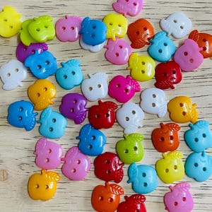 Könnte beinhalten: Eine Sammlung von bunten Knöpfen in Apfelform in verschiedenen Farbtönen, darunter Gelb, Grün, Blau, Rosa, Rot, Orange, Weiß und Lila. Jeder Knopf hat zwei Löcher zum Annähen und ein Blattdetail. Die Knöpfe sind auf einer hellen Holzoberfläche verstreut.