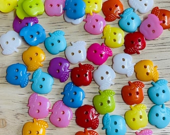 May include: A collection of colorful apple-shaped buttons in various shades, including yellow, green, blue, pink, red, orange, white, and purple. Each button has two holes for sewing and a leaf detail. The buttons are scattered on a light wooden surface.