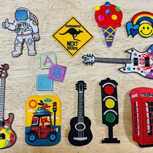May include: A selection of embroidered patches with diverse designs. Includes an astronaut, a kangaroo road sign, an ice cream cone, a rainbow with a smiley face, guitars, ABC blocks, a car with dogs, a traffic light, and a red telephone box.