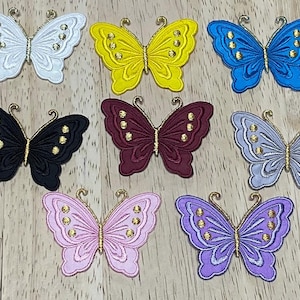 May include: Ten iron-on butterfly patches in various colours, including black, white, yellow, blue, purple, pink, and maroon. Each butterfly has gold accents and a detailed design.