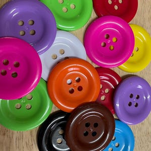May include: A collection of round, four-hole plastic buttons in a variety of vibrant colours including lime green, red, purple, magenta, yellow, orange, white, brown, black, and blue, arranged in a scattered, overlapping pattern on a wooden surface.