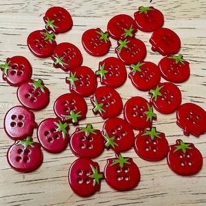 May include: A collection of small, round, red buttons shaped like tomatoes. Each button features four central holes and a green painted stem detail at the top. The buttons are scattered across the frame, displaying a glossy finish and uniform design.