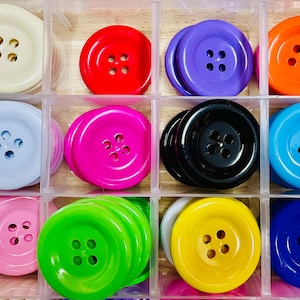 May include: A selection of colourful, round buttons in a clear plastic organiser. The buttons are in shades of red, purple, orange, blue, green, pink, yellow, black, and white. Each button has four holes for sewing.