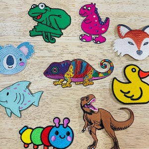 May include: A set of ten colorful iron-on patches featuring various animals and designs. The patches include a frog, a dinosaur, a fox, a koala, a chameleon, a duck, a shark, a dinosaur, a caterpillar, and a T-Rex.