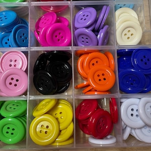May include: A clear plastic storage container with twelve compartments, each filled with a collection of round, four-hole plastic buttons in various solid colours including blue, pink, purple, cream, black, orange, green, yellow, red, and white.