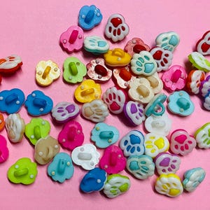 Könnte beinhalten: Eine Sammlung bunter Pfotenabdruck-Knöpfe in verschiedenen Farben wie Blau, Rosa, Gelb und Grün. Die Knöpfe haben ein Zwei-Loch-Design und sind auf einer rosa Oberfläche verstreut. Ideal für Bastelprojekte.