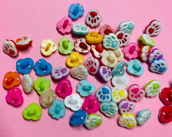 May include: A collection of colorful paw print-shaped buttons in various colors, including blue, pink, yellow, and green. The buttons have a two-hole design and are scattered on a pink surface. These decorative buttons are ideal for craft projects.