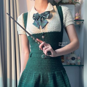 Peut inclure: Robe verte en crochet avec un haut en crochet blanc et un nœud papillon vert. La robe a une jupe plissée et une ceinture. Une baguette est tenue dans la main. Le texte "moon & baileys" est visible.