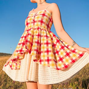 Puede incluir: Un vestido de verano con un corpiño a cuadros rojos, amarillos y blancos y tirantes finos. La falda tiene un diseño en capas, con un ribete de ganchillo en el dobladillo. El vestido se usa al aire libre sobre un fondo de hierba alta y cielo azul claro.