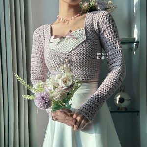 Puede incluir: Top de crochet lila claro con mangas largas y cuello cuadrado. El top se combina con una falda blanca. La imagen también presenta un ramo de flores y un collar. El texto "moon & baileys" es visible.