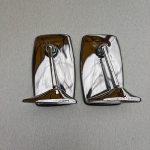 Puede incluir: Dos espejos retrovisores laterales rectangulares cromados. Cada espejo tiene una superficie reflectante curvada y un brazo largo y delgado que se conecta a una base. Los espejos están diseñados para un vehículo y están sobre un fondo neutro.