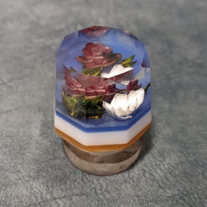 Puede incluir: Objeto decorativo de resina con una parte superior facetada, que muestra rosas rojas y flores blancas incrustadas. El objeto tiene un diseño en capas azul, blanco y naranja, colocado sobre una base gris. El arreglo floral está encapsulado en resina transparente.