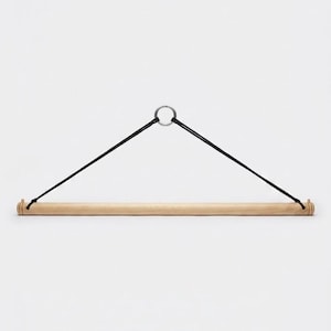 Op de afbeelding: Een lichtgekleurde houten roede ontworpen om op te hangen, voorzien van een zwart koord aan elk uiteinde dat samenkomt bij een centrale zilverkleurige metalen ring. De houten roede heeft kleine eindkapjes en een gladde, cilindrische afwerking.