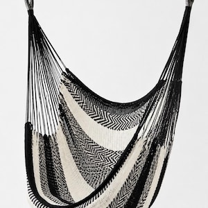 Puede incluir: Una silla colgante con un asiento de tela tejida con un patrón de espiga en blanco y negro. La silla está suspendida por cuerdas negras unidas a mosquetones de metal en la parte superior, creando una estructura de asiento profunda y curva.