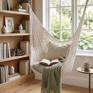 Puede incluir: Una silla hamaca de cuerda tejida color crema suspendida del techo, con un cojín cuadrado beige, un libro abierto sobre el asiento y una manta de color verde salvia con flecos colocada sobre el borde delantero del asiento.