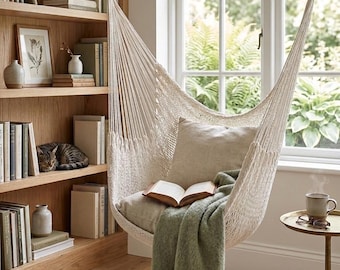 May include: A cream-colored woven rope hammock chair suspended from the ceiling, featuring a square beige cushion, an open book resting on the seat, and a sage green fringed throw blanket draped over the front edge of the seat.