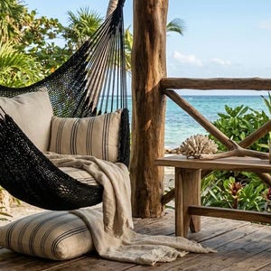 Puede incluir: Una silla hamaca tejida en color negro suspendida de un poste de madera, con un cojín beige de rayas negras finas, una manta a juego y un cojín de suelo a rayas, situada en una terraza de madera con vistas al océano.