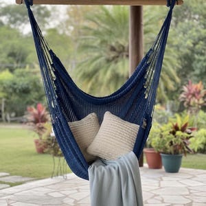 May include: A navy blue woven hammock chair suspended from a horizontal wooden spreader bar. The chair features a deep seat with two square beige textured cushions and a light grey fringed throw blanket draped over the front edge.