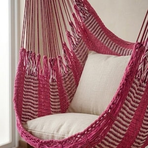 Puede incluir: Una silla colgante con un diseño tejido en tonos rosa y blanco. La silla tiene un cojín suave de color crema en el interior. La silla está suspendida por múltiples cuerdas rosas, creando un espacio acogedor y acogedor para la relajación.