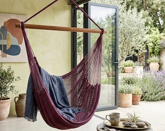 May include: A hanging hammock chair in a deep burgundy color, suspended from a wooden beam. A blue blanket rests inside the chair. The chair hangs in front of a glass door, with a view of a garden.