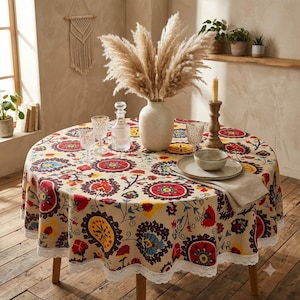 Könnte beinhalten: Eine runde Tischdecke mit cremefarbenem Hintergrund und einem bunten Blumen- und Kreismuster in Rot, Gelb und Dunkelbraun. Der Rand ist mit weißer Spitzenborte verziert. Der Tisch ist mit einer Glaskaraffe, zwei Gläsern, einer Schale und einer Vase gedeckt.