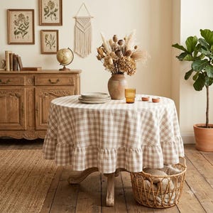 Peut inclure: Une table ronde recouverte d'une nappe à carreaux beige et blanc avec un bord à volants. Sur la table se trouvent un vase en céramique rempli de fleurs séchées, une pile d'assiettes et un petit verre ambré. Un panier tressé repose au sol.