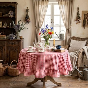 May include: A round table covered with a red and white gingham tablecloth featuring a ruffled edge. On the table sits a floral porcelain teapot, matching teacups and saucers, a small creamer, a book, and a glass vase filled with colorful flowers.