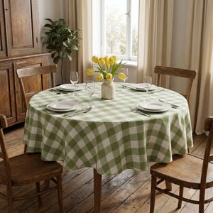 Peut inclure: Une table à manger ronde recouverte d'une nappe à carreaux vert sauge et blanc. La table est dressée avec quatre assiettes blanches, quatre verres à vin transparents et des couverts en argent. Un vase en céramique avec des tulipes jaunes.