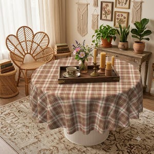May include: A round table covered with a brown, white, and green plaid tablecloth. On the table sits a dark wooden rectangular tray holding a ceramic cup, a glass vase with colorful flowers, and three cylindrical yellow candles of varying heights.