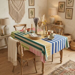 Op de afbeelding: Een rechthoekig tafelkleed met een levendig patroon van verticale strepen in blauw-, geel-, groen- en roze tinten. De stof ligt over een houten eettafel, die is gedekt met keramische kommen, mokken en glazen vazen.