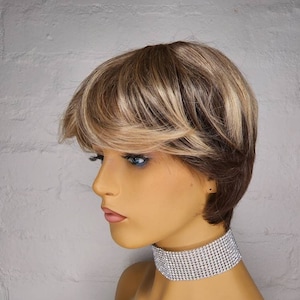 May include: A short, layered wig with brown and blonde highlights. The wig has a side part and is styled with a fringe. The wig is worn by a mannequin with a silver rhinestone choker.