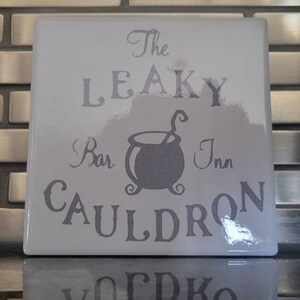 Puede incluir: Una baldosa cuadrada blanca con las palabras "The Leaky Bar Inn Cauldron" en una fuente estilizada. Un gráfico de caldero está centrado en la baldosa. La baldosa se refleja en una superficie brillante.