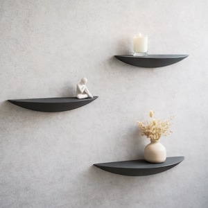 Op de afbeelding: Drie donkergrijze, halfronde zwevende planken aan een lichtgrijze muur. De planken bevatten een witte kaars, een klein wit beeldje en een kleine vaas met gedroogde bloemen. Een modern woondecoratie-item.