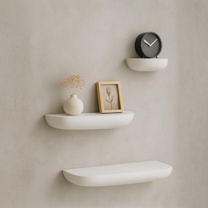 May include: Three white, semi-circular floating shelves mounted on a light gray wall. The top shelf holds a black clock with white hands. The middle shelf displays a small vase with dried flowers and a framed picture. The bottom shelf is empty.