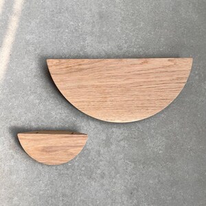 K&ouml;nnte beinhalten: Zwei halbkreisf&ouml;rmige Holzregale, eines gr&ouml;&szlig;er als das andere, sind vor einem grauen Betonhintergrund zu sehen. Die Regale sind aus hellbraunem Holz mit nat&uuml;rlicher Maserung.