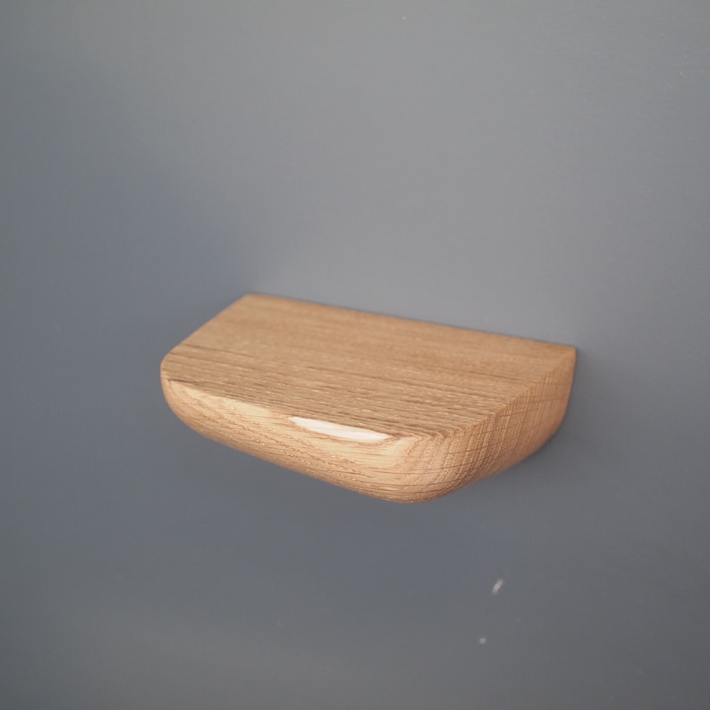 K&ouml;nnte beinhalten: Ein kleines, halbkreisf&ouml;rmiges Wandregal aus hellem Holz mit sichtbarer nat&uuml;rlicher Maserung. Das Regal verf&uuml;gt &uuml;ber eine flache Oberseite und eine abgerundete, geschwungene Vorderkante, die von einer glatten vertikalen Fl&auml;che absteht.