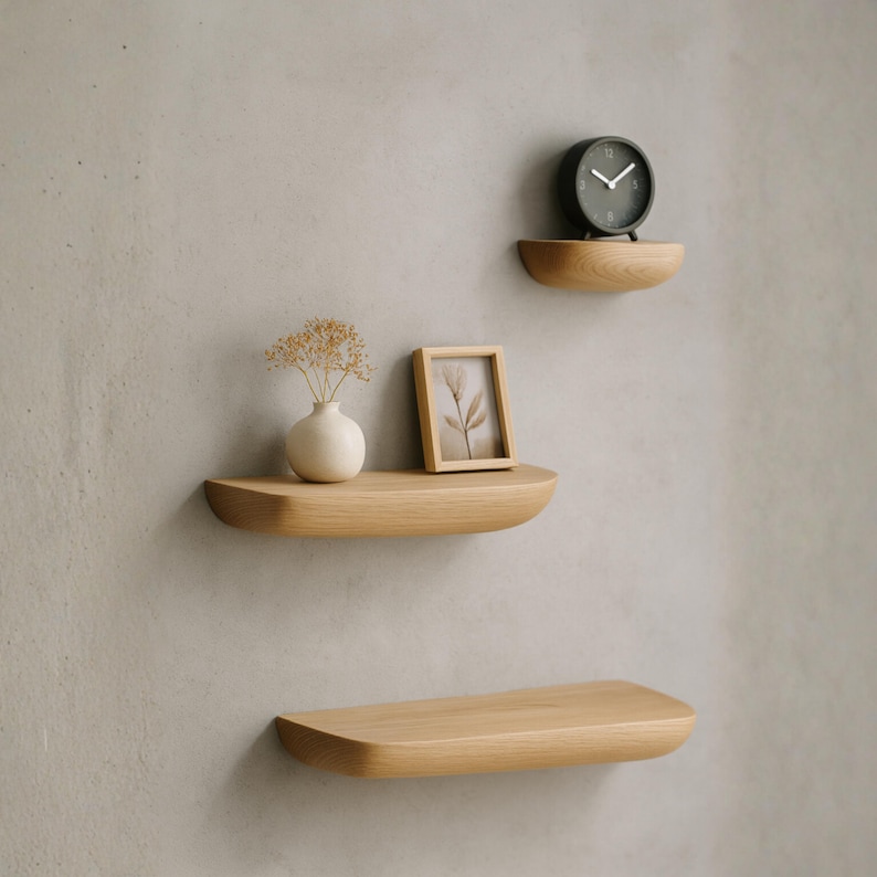 K&ouml;nnte beinhalten: Drei halbkreisf&ouml;rmige, an der Wand montierte Wandregale aus hellem Holz. Das oberste Regal tr&auml;gt eine kleine schwarze Analog-Uhr. Das mittlere Regal enth&auml;lt eine wei&szlig;e Keramikvase mit Trockenblumen und einen kleinen Bilderrahmen aus Holz.