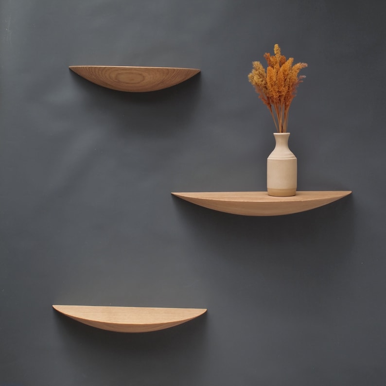 May include: Three wooden floating shelves with a curved design are mounted on a gray wall. The shelves are made of light-colored wood. A small, off-white vase with dried flowers sits on the middle shelf, adding a touch of natural decor.
