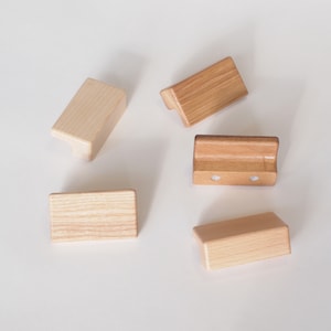 May include: Five wooden cabinet pulls in various shapes. The pulls are made of light-coloured wood with visible grain patterns. Some have a rectangular shape, while others have a curved or angled design. The pulls are scattered on a white surface.