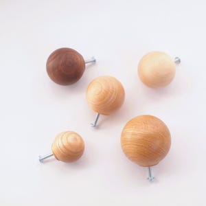 May include: Five wooden ball knobs in various shades of brown and tan, each with a silver screw. The knobs are arranged on a white background. The wood grain is visible on some of the knobs.